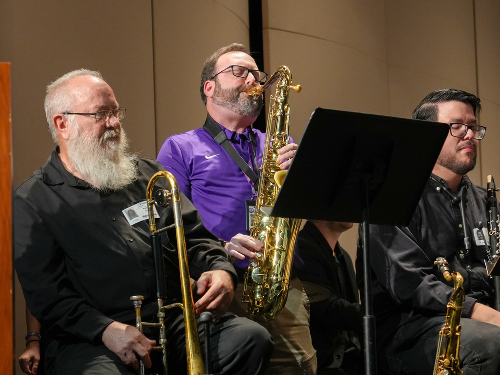 Four men in black shirts and glasses playing instruments, including saxophones and trombones, seated on stage with a music stand in the background.