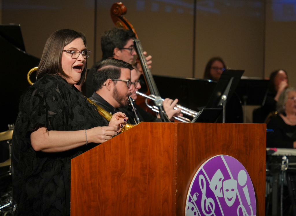 A woman singing at a podium with a trumpet player. Behind her, a band with cello, piano, and drums plays.