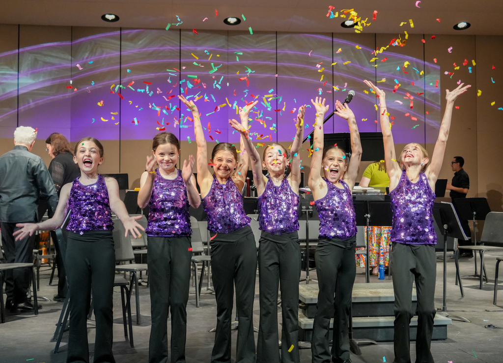 Five young girls in purple sequined tops and black pants stand on a stage with hands raised. Confetti falls around them.