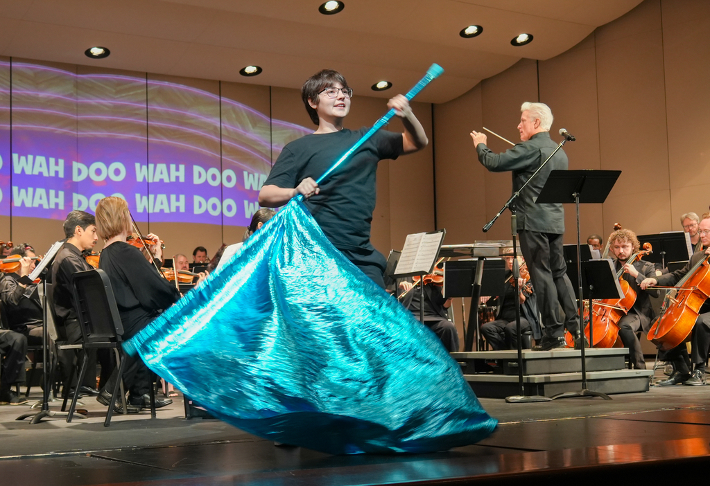 Performer on stage with a blue fabric, holding a blue staff, with orchestra behind, spotlights overhead.