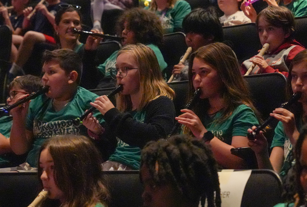 A group of children playing flutes seated in rows, some wearing green shirts, others not.