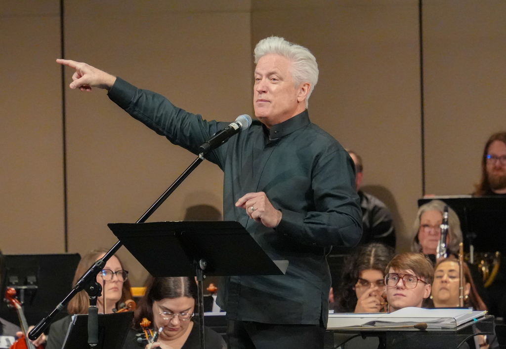A man stands in front of a music stand, pointing while speaking. A band sits behind him.