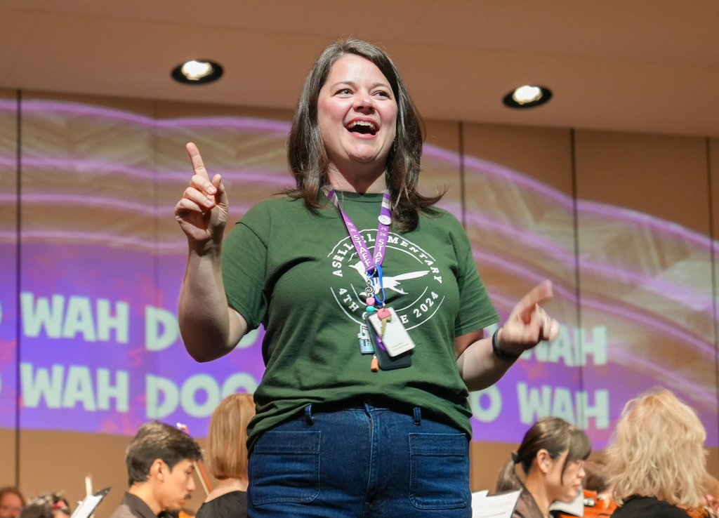 A woman in a green shirt and blue jeans gestures on stage with a purple lanyard around her neck.