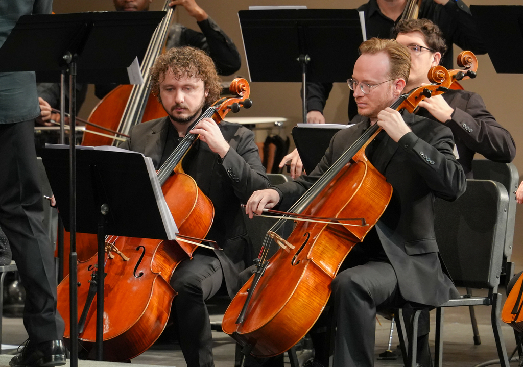 Musicians sit on chairs in a concert hall, playing cellos. Some people in black sit behind them. Sheet music stands are in front of the musicians.
