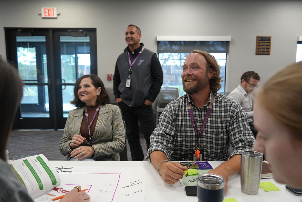 A group of people are in a meeting room. Some are seated, others are standing. A man in a plaid shirt smiles.