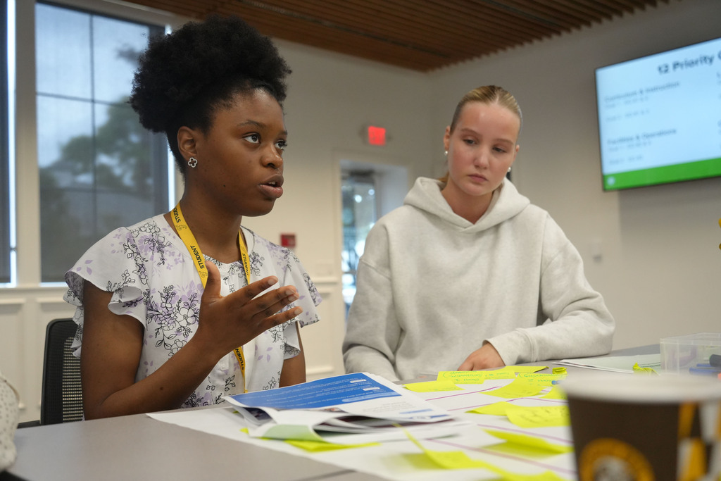 Two women in a classroom setting with one speaking and the other listening, surrounded by open notebooks, sticky notes, and a cup.