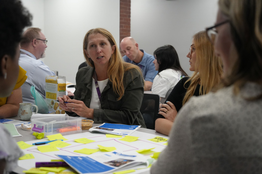 Several people in a meeting room with a woman speaking while others listen, tables with sticky notes and papers.
