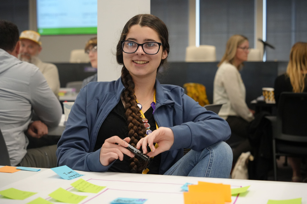 A woman wearing glasses sits at a desk, smiling, surrounded by coworkers. Papers with sticky notes are on the table.