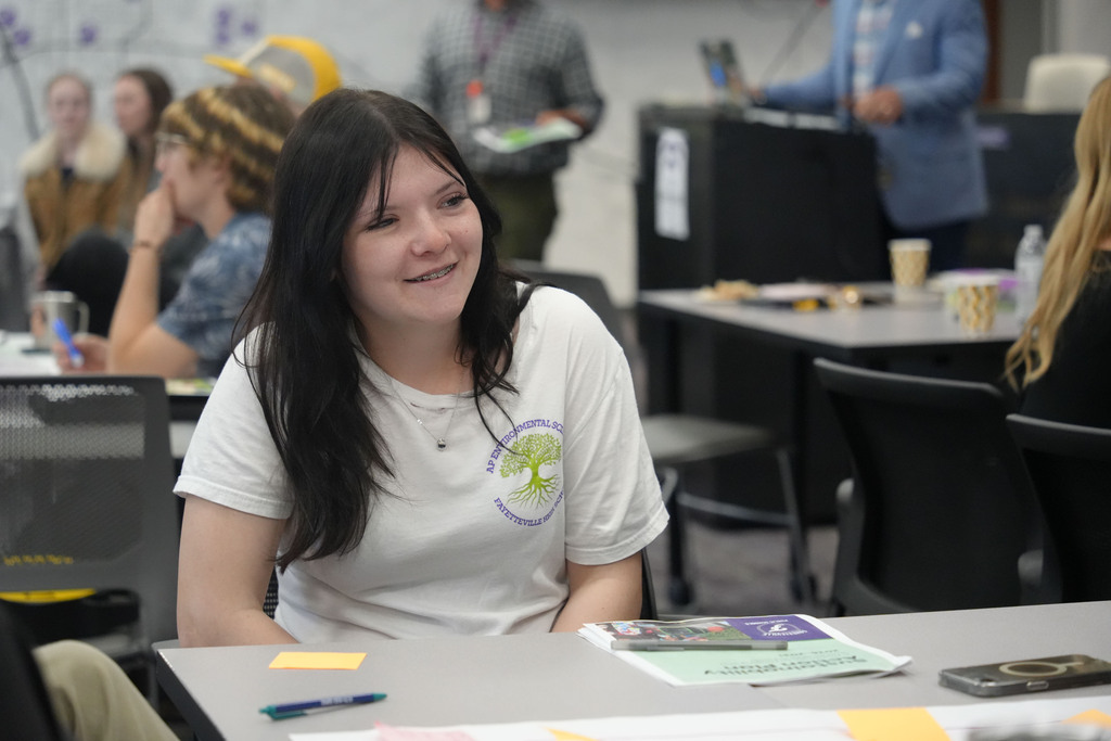 A woman with long dark hair smiles while seated at a table with papers, surrounded by others in a room.