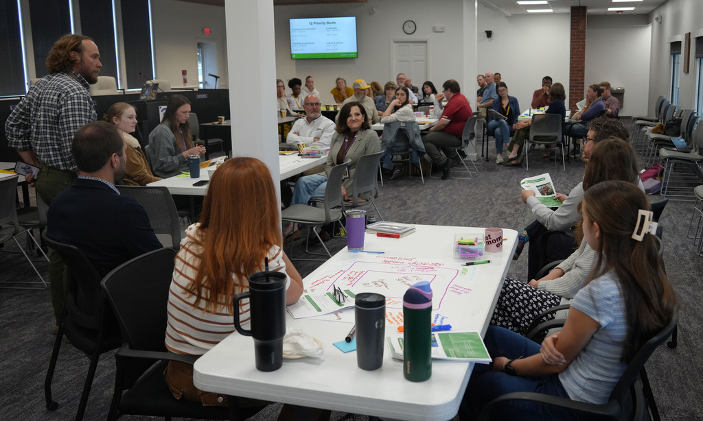 A room with multiple tables, people seated around them, a projector screen, and a floor map.