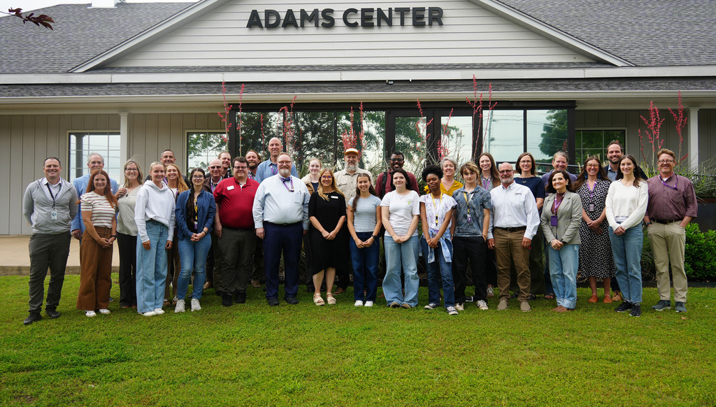 Group of people standing in front of the Adams Center, wearing casual clothing, on a grassy lawn.