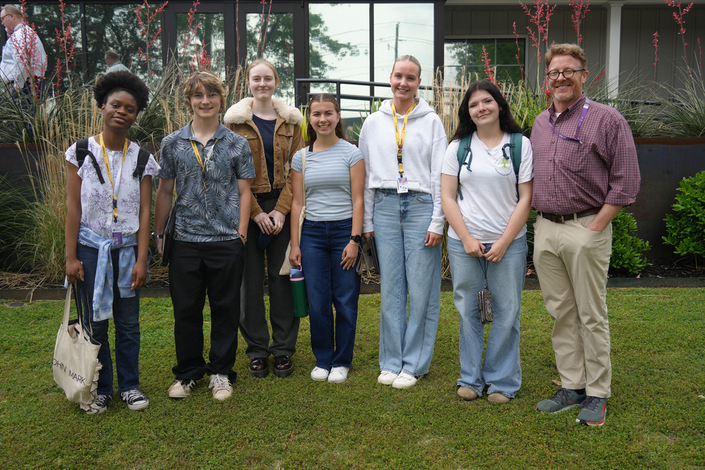 Eight individuals, likely students, pose for a photo on a lawn, wearing casual attire. The background shows a building with greenery.