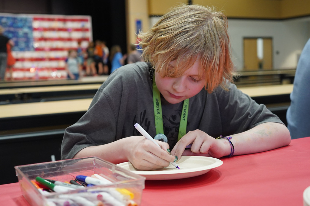 A child with blond hair draws on a paper plate using a marker. Supplies are in a clear container.