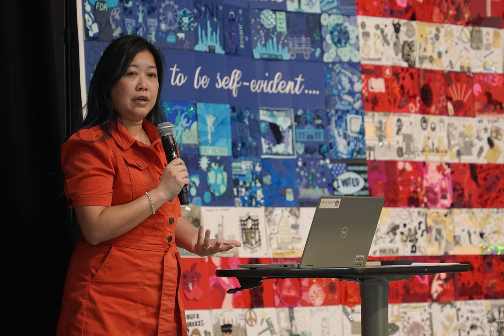Woman in orange dress speaking into a microphone in front of an American flag backdrop.