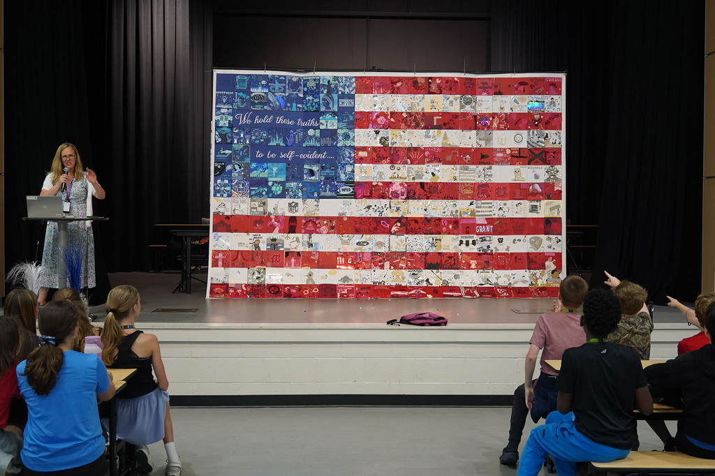 A woman at a podium speaks to children seated in front of an American flag made of pieces of fabric.