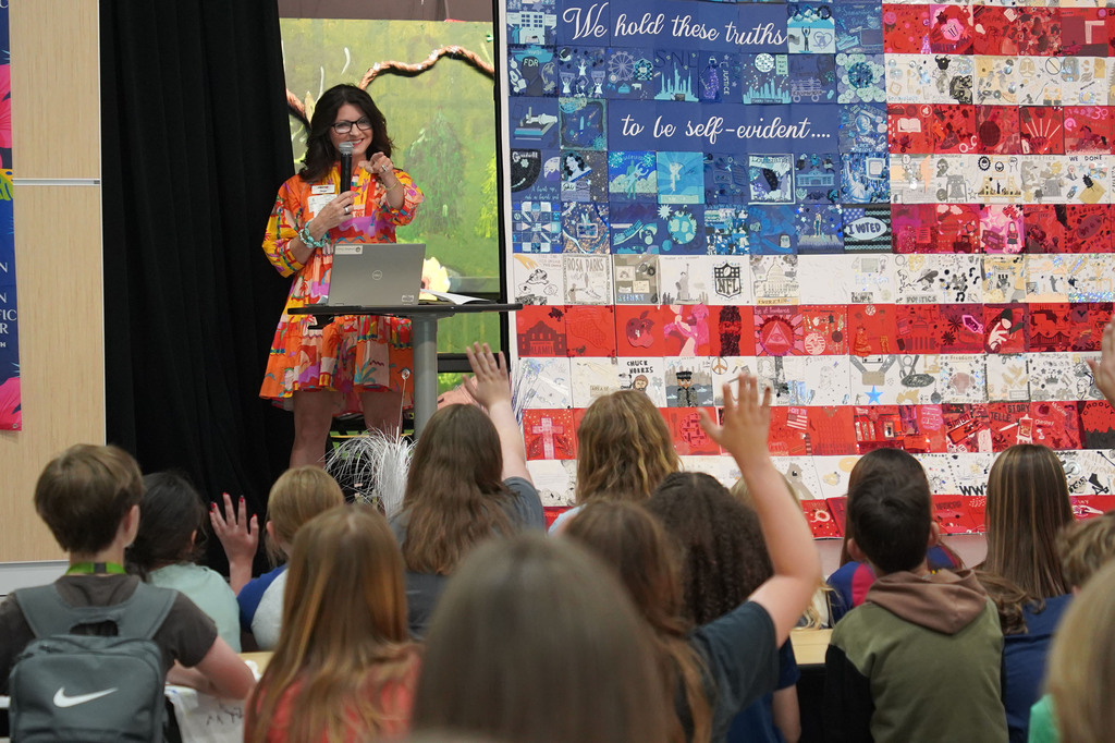 A woman stands at a podium before an American flag mural, speaking to a classroom full of students.