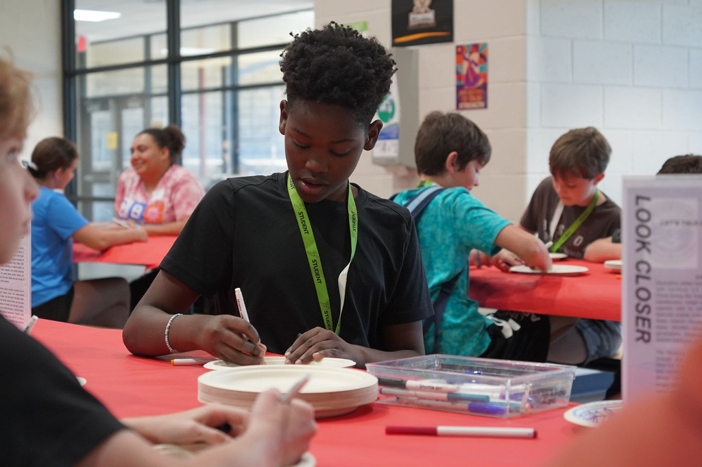 A group of students seated around tables, one boy drawing on a paper plate. Red tablecloths.