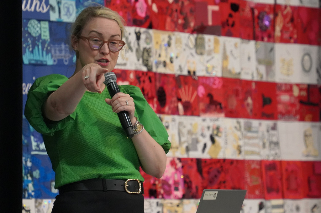 A woman wearing a green blouse and glasses speaks into a microphone before a collage backdrop.