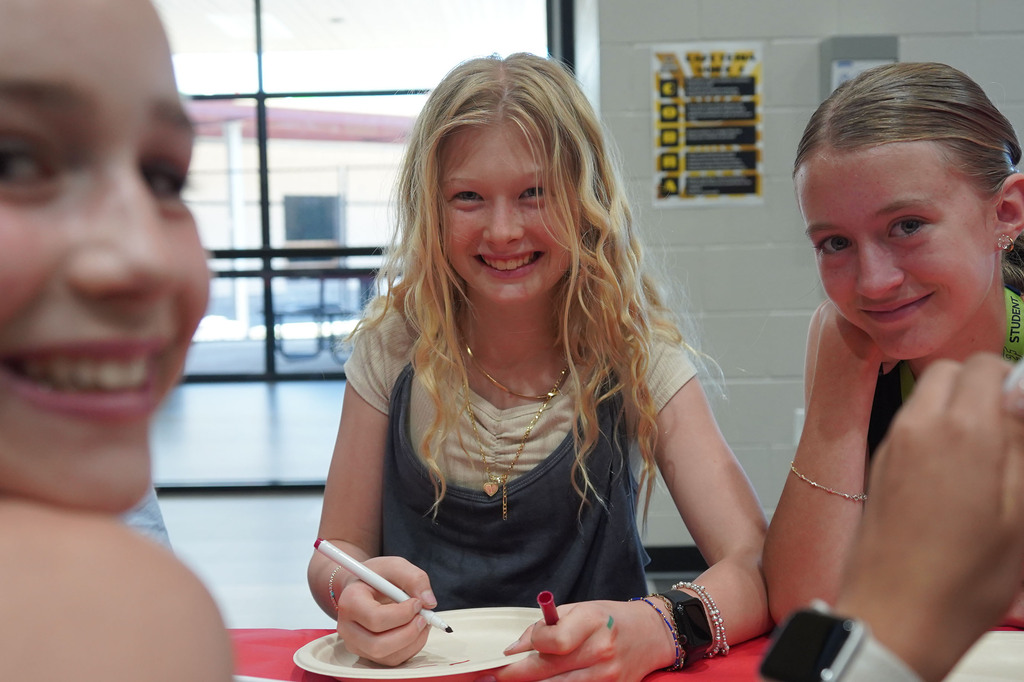 Three young girls smiling and sitting at a table. One girl is holding a pen and paper.