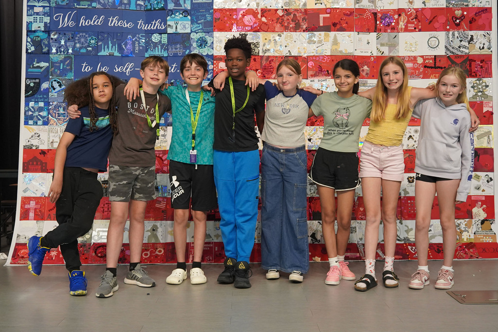 Seven kids in casual clothing pose with an adult in front of a colorful flag mural.
