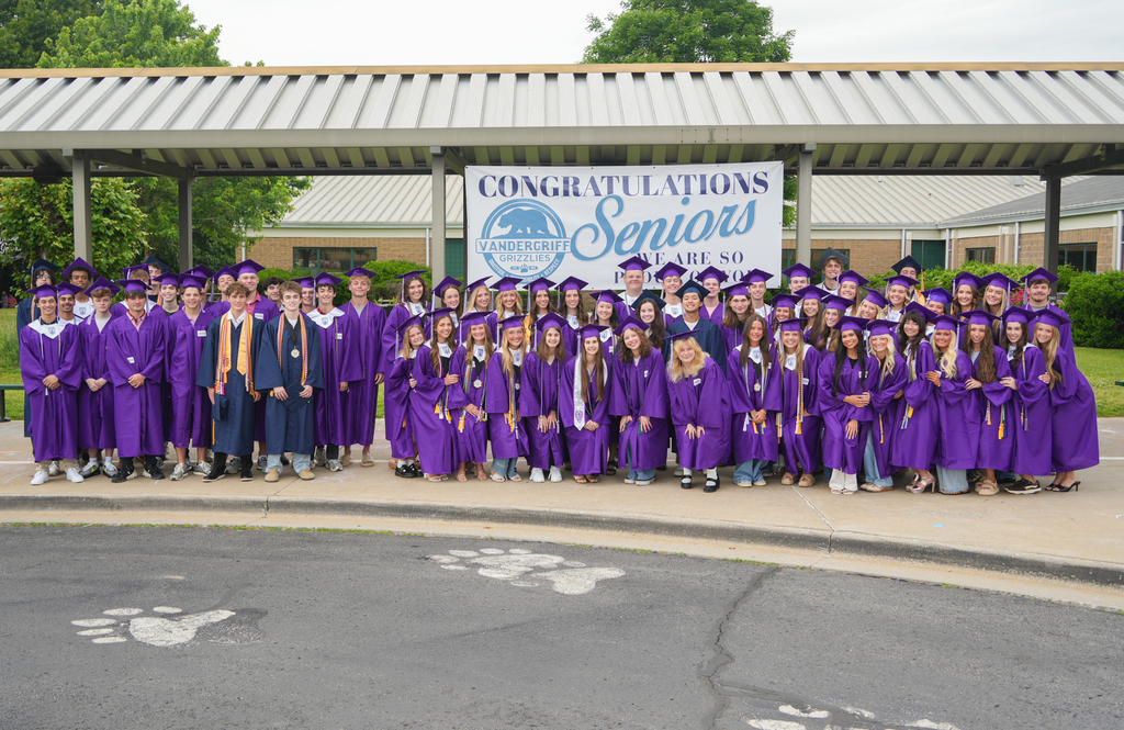 A group of people in purple gowns and caps stand under a banner reading "Congratulations Seniors." The background shows trees and a building.