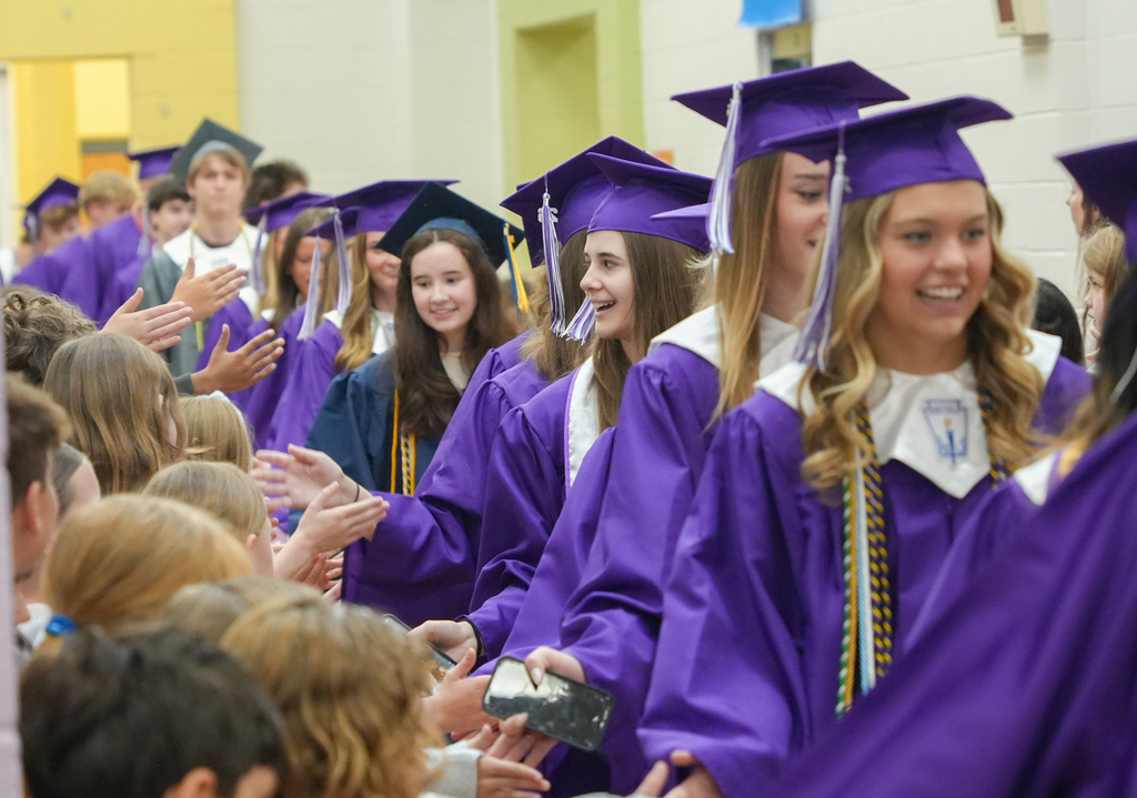 Graduates in purple gowns and caps at a school event. Some are smiling and waving.