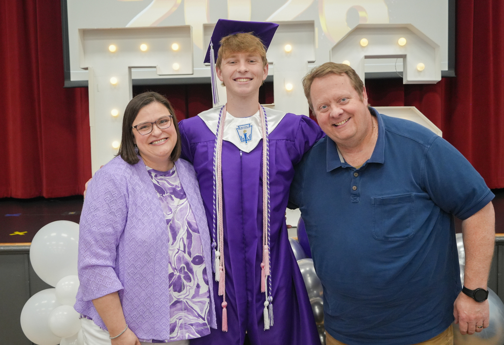 Three people pose for a photo with balloons in the background. A graduate in a cap and gown stands between a woman and a man.