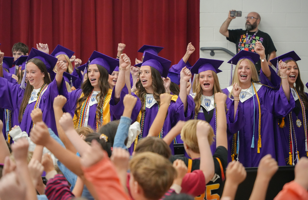 A group of people in purple gowns and hats, raising their fists, sing in front of an audience.
