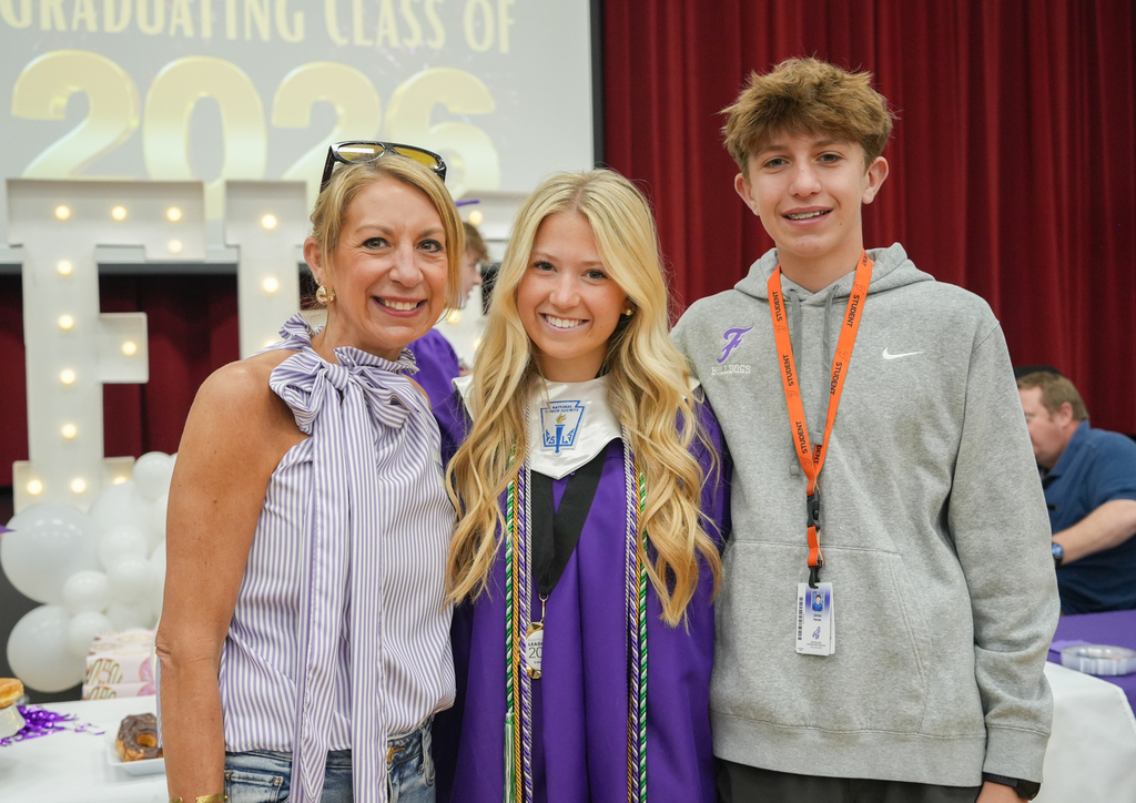 Three people stand before a red curtain with "GRADUATING CLASS OF 2026" written above. A woman and a woman in a cap and gown smile.
