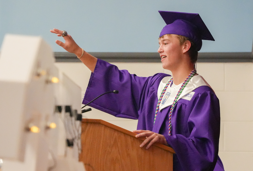 A person in a purple graduation gown, holding a microphone, and giving a speech at a podium.