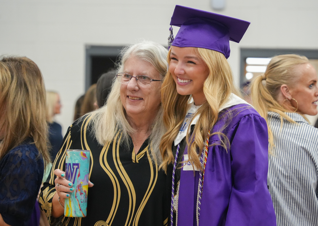 Two women smile in a room with a white wall. One wears a purple cap and gown, the other holds a cup.
