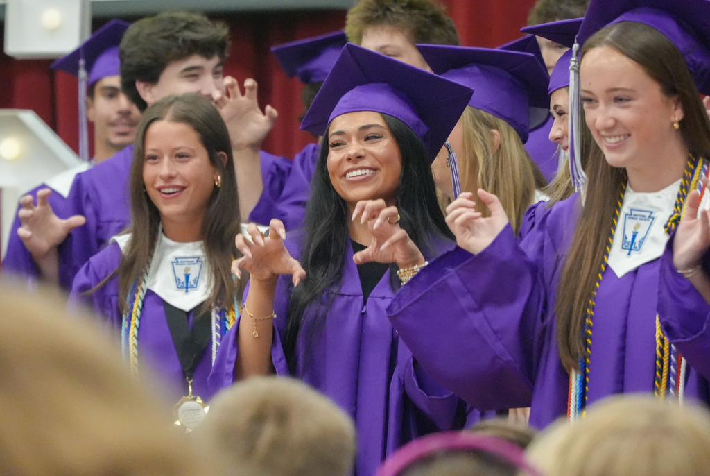 Graduates in purple gowns and caps clap for their peers in a room with red curtains.