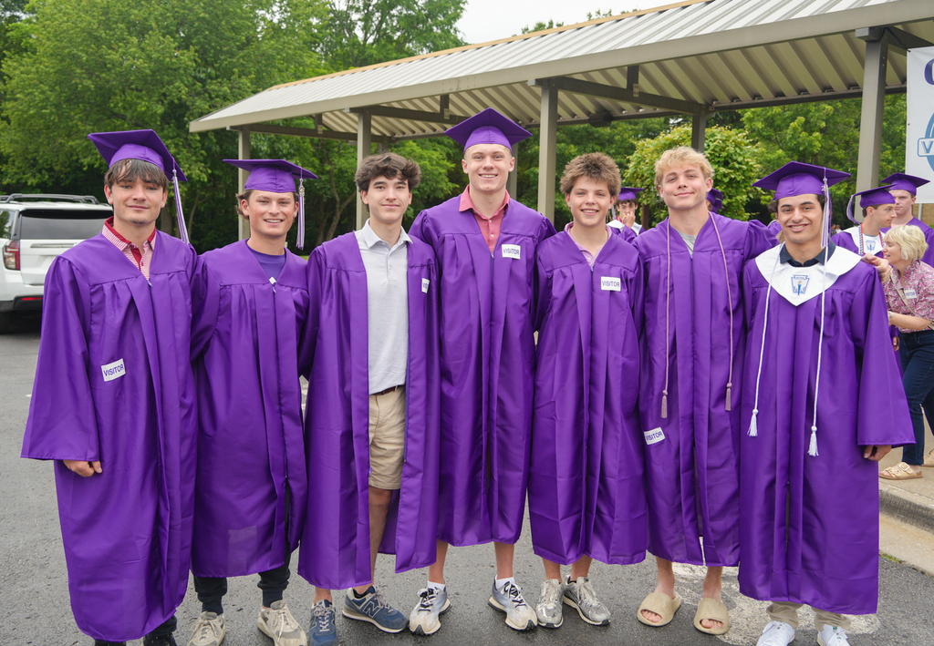 Seven people stand in purple graduation gowns; one wears a white shirt. The background includes trees and a shelter.