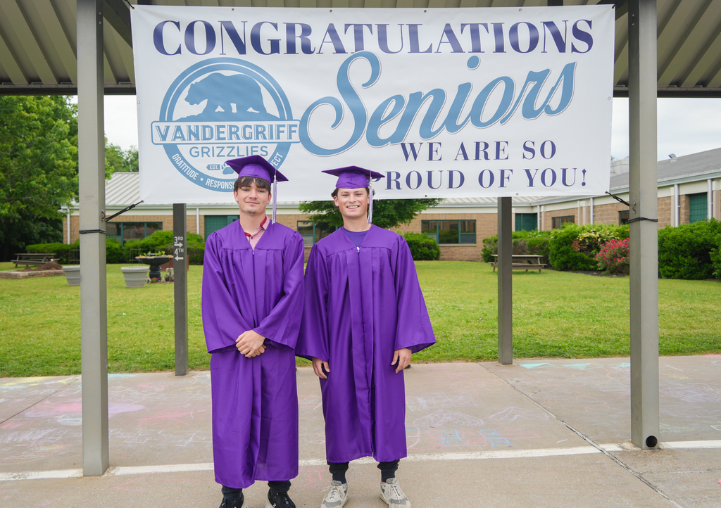 Two students in purple graduation gowns stand in front of a sign that reads "Congratulations Seniors".