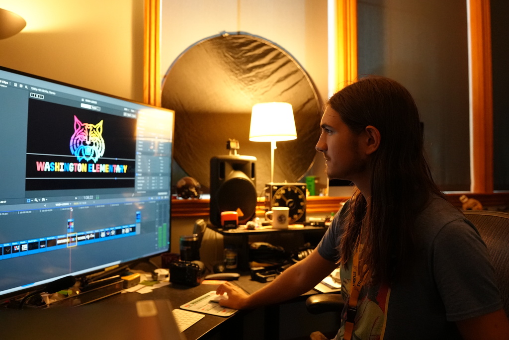 A person with long hair sits at a desk in front of a computer screen.