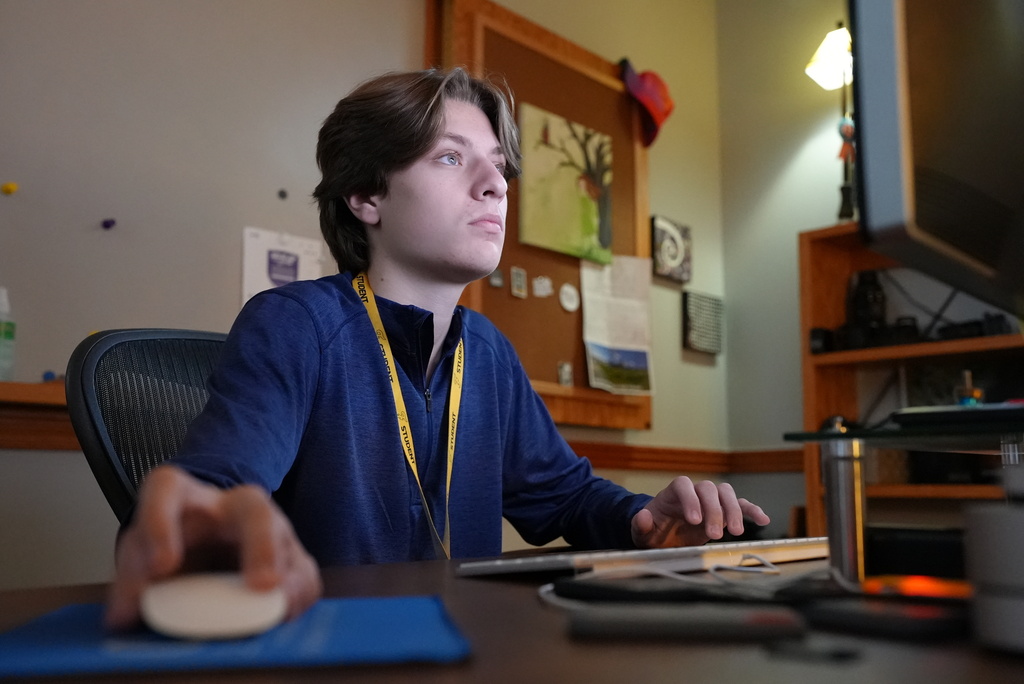 Person seated, focused on computer, using a mouse on a desk, surrounded by a bulletin board and shelves.