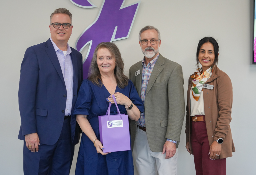 Five people stand together, smiling. A woman in a blue dress holds a purple bag. The wall behind them has a purple design.