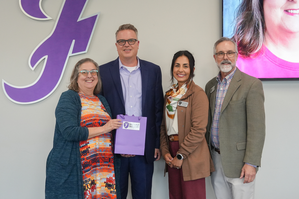 Four people stand in front of a wall with a purple F logo. They hold a purple bag.