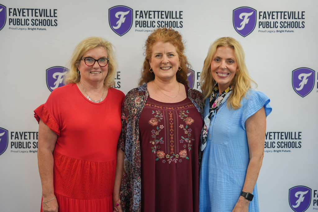 Three women pose for a picture in front of a wall with the text "Fayetteville Public Schools". They are wearing red, maroon, and blue clothing.