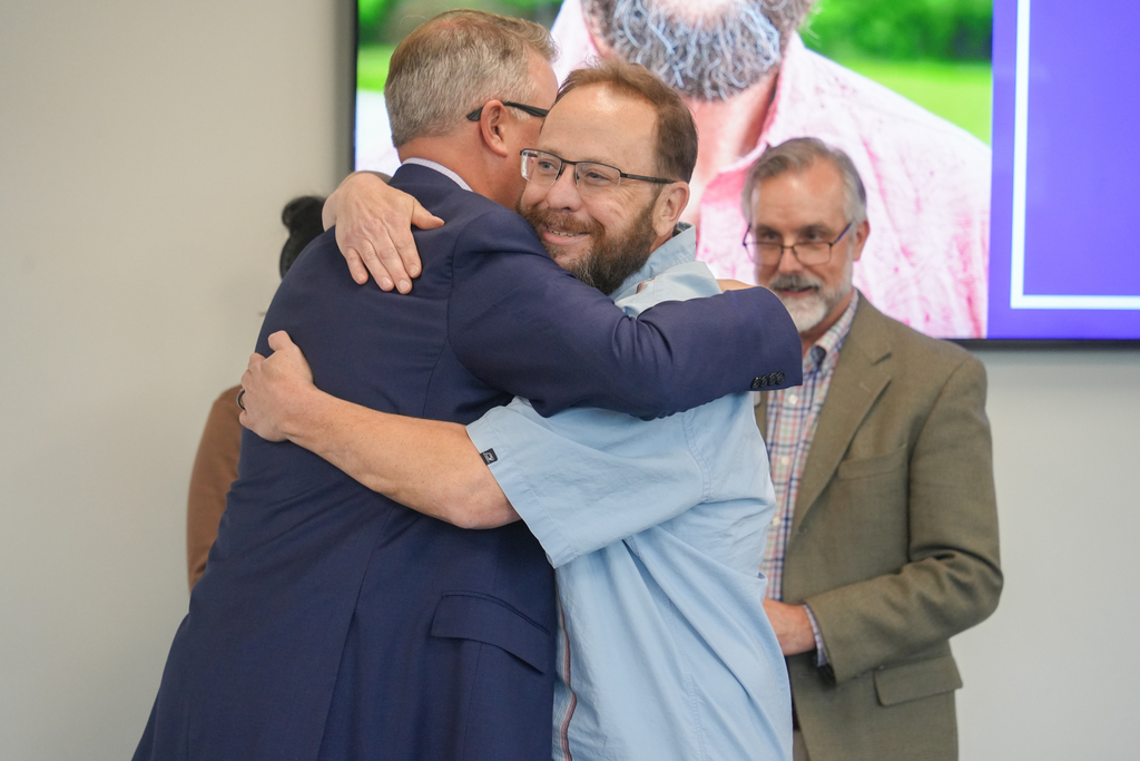 Two men hug in a room. A man in glasses stands to the side, watching. A screen displays a photo of a bearded man.