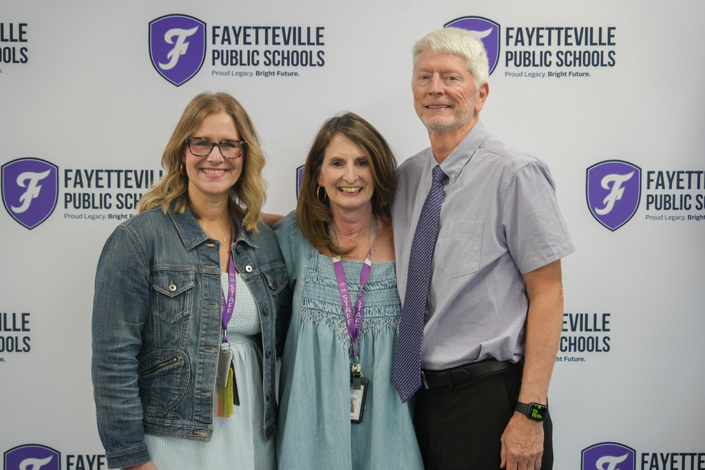 Three people pose for a photo, standing together in front of a wall with the words "Fayetteville Public Schools."