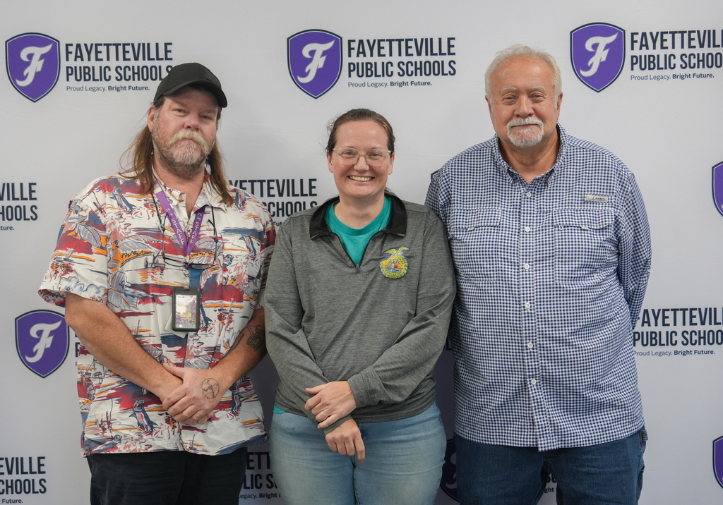 Three people stand in front of a wall with "Fayetteville Public Schools" logos. The man on the left wears a cap and a colorful shirt.