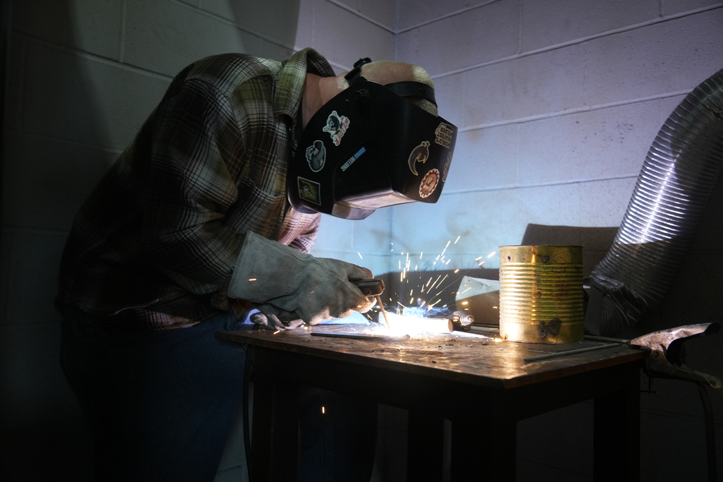 A person in a welding mask works at a table, sparks fly as they weld metal.