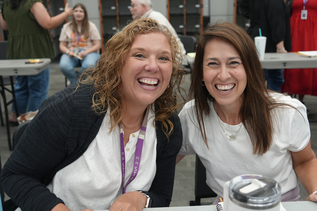 Two women, one with curly hair and the other with straight hair, smiling and sitting at a table.