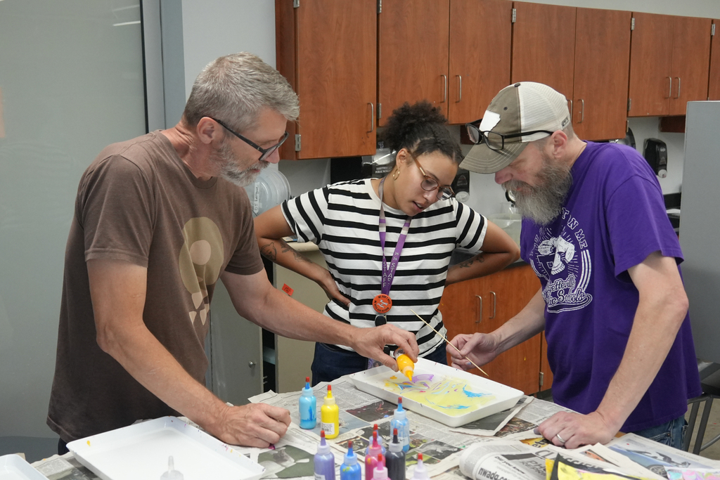 Three adults with protective coverings work on a paper with paint and paper. Wooden cabinets in the background.