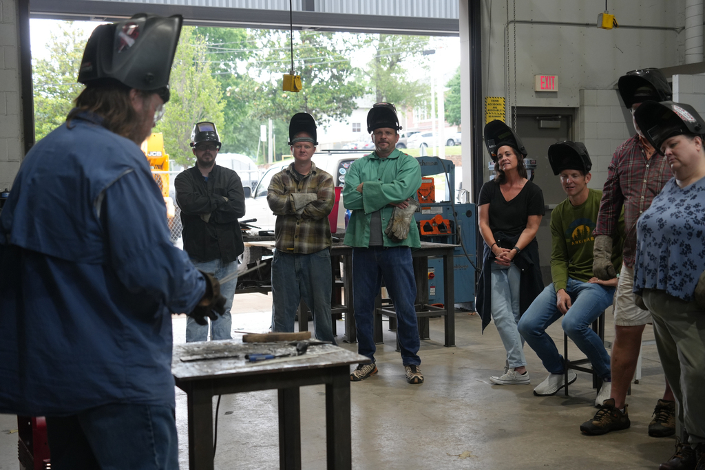A workshop with a man in blue leading a group. Participants wear welding helmets and jackets, standing around a table.