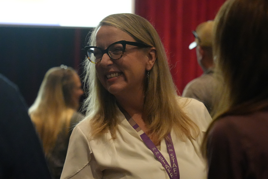 Woman with blond hair and glasses smiles. She wears a white shirt with a purple lanyard. Behind her, a man with a brown shirt and a woman with blond hair.