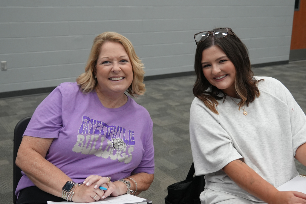 Two women sit next to each other. The woman on the left wears a purple shirt with writing on it.