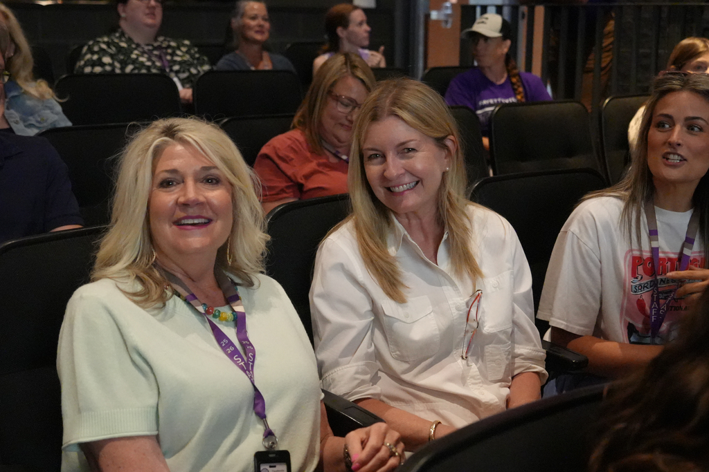 A group of women sit in a theater, smiling. Two women in white shirts sit close together.