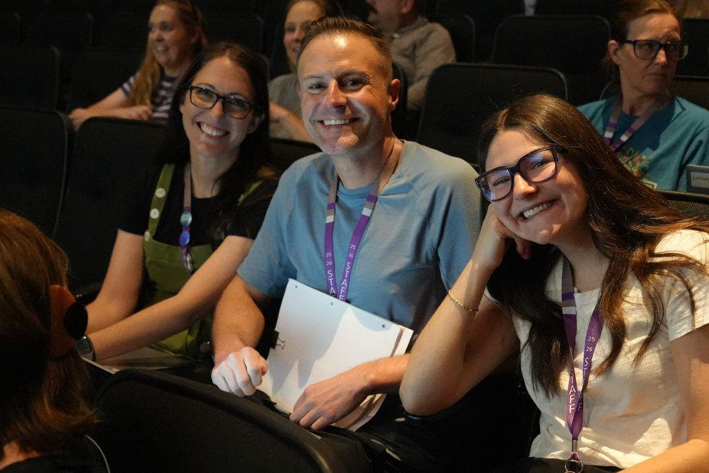 Four people sit in a row of theater seats; two women wear glasses and IDs, and a man holds a folder.
